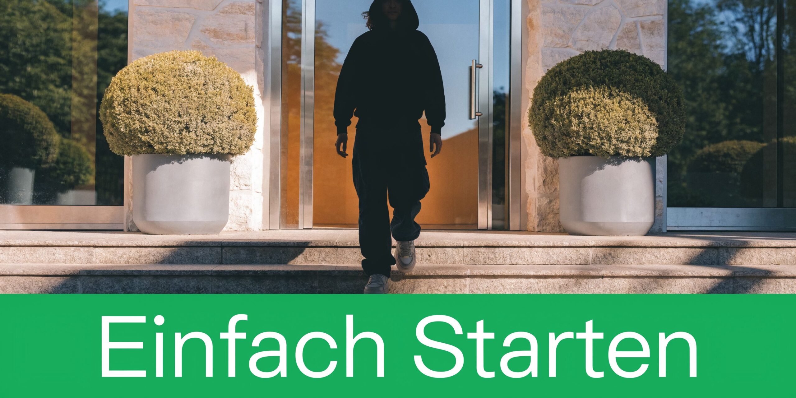 Eine Person in einem schwarzen Kapuzenpullover verlässt ein modernes Gebäude mit zwei großen Pflanzen vor dem Eingang.