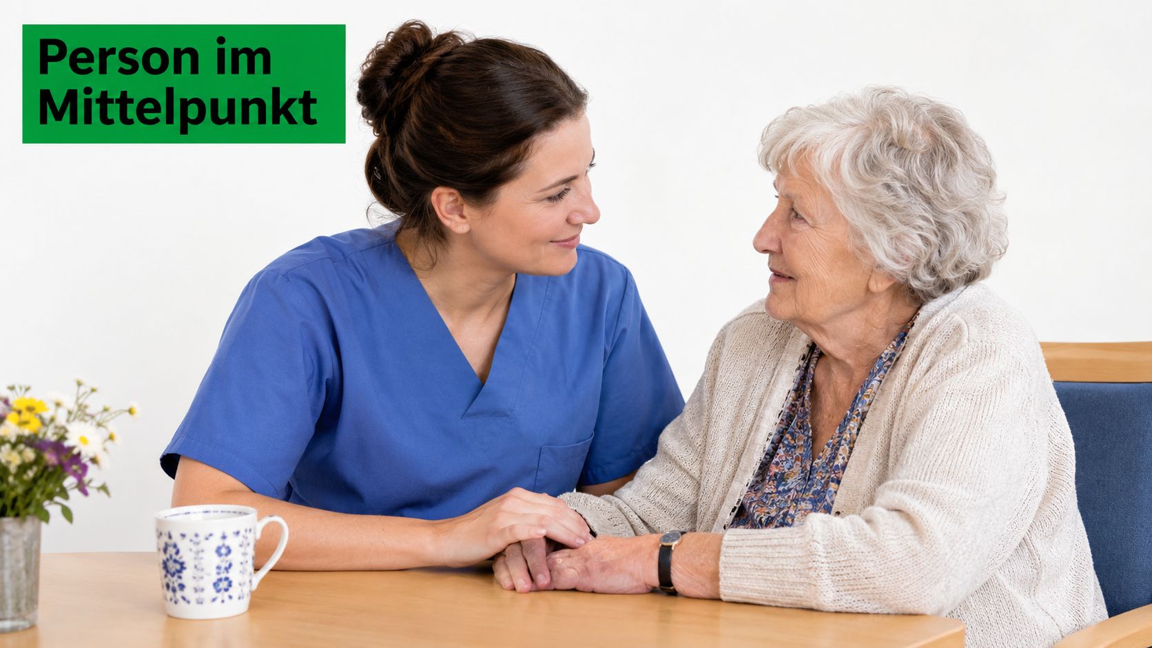 Eine freundliche Pflegekraft hält die Hand einer älteren Dame, die an einem Tisch in einem Gespräch sitzt.